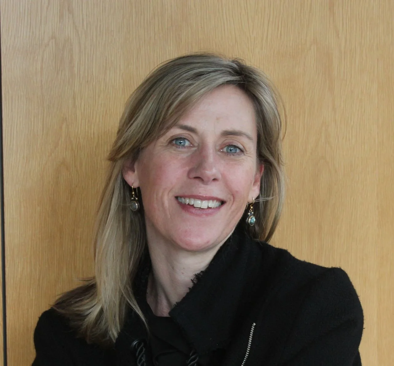 A smiling woman with blonde hair, blue eyes, wearing earrings and a black jacket, standing in front of a wooden background.