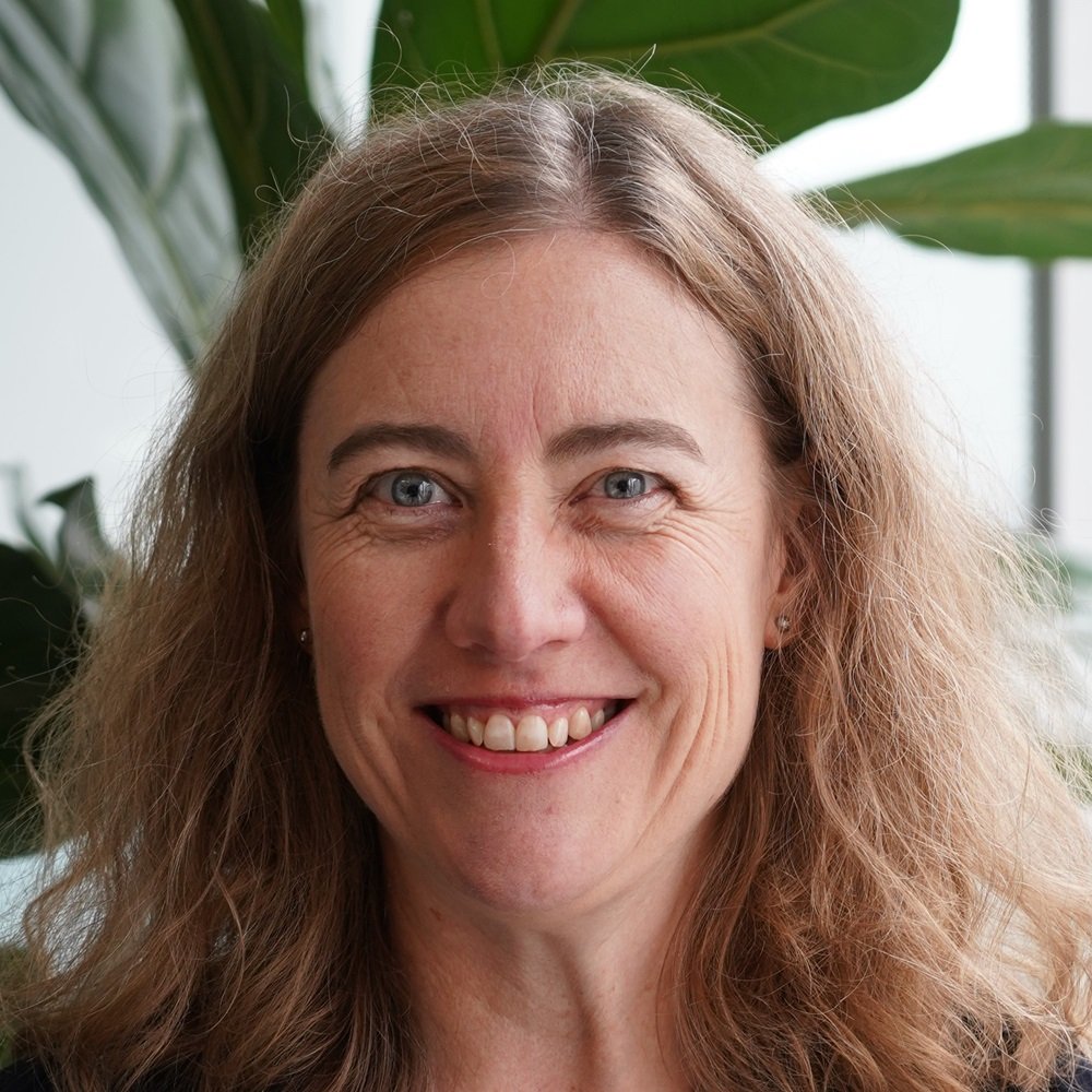 Close-up of a smiling woman with blue eyes and wavy light brown hair, standing indoors with green plants in the background.