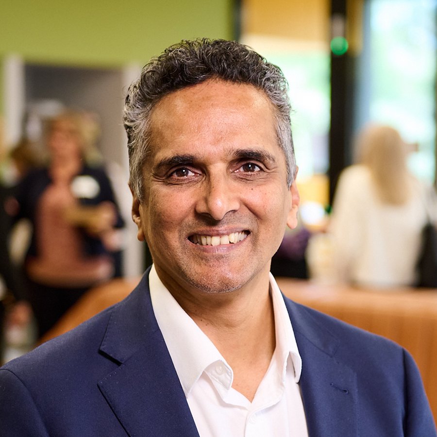 A man smiling in a professional setting with blurred people and bright lighting in the background.