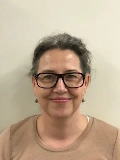 A woman with short gray hair and glasses smiling at the camera, wearing a beige top, against a plain light-colored background.