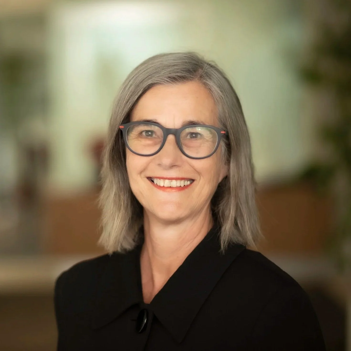 A middle-aged woman with gray hair, wearing glasses and a black top, smiling at the camera with a blurred indoor background.