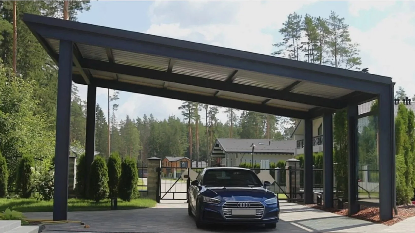 A modern metal steel carport with a metal roof and support beams, sheltering a blue Audi sedan parked underneath, surrounded by manicured bushes and a driveway leading to metal gates, with a property and trees in the background.