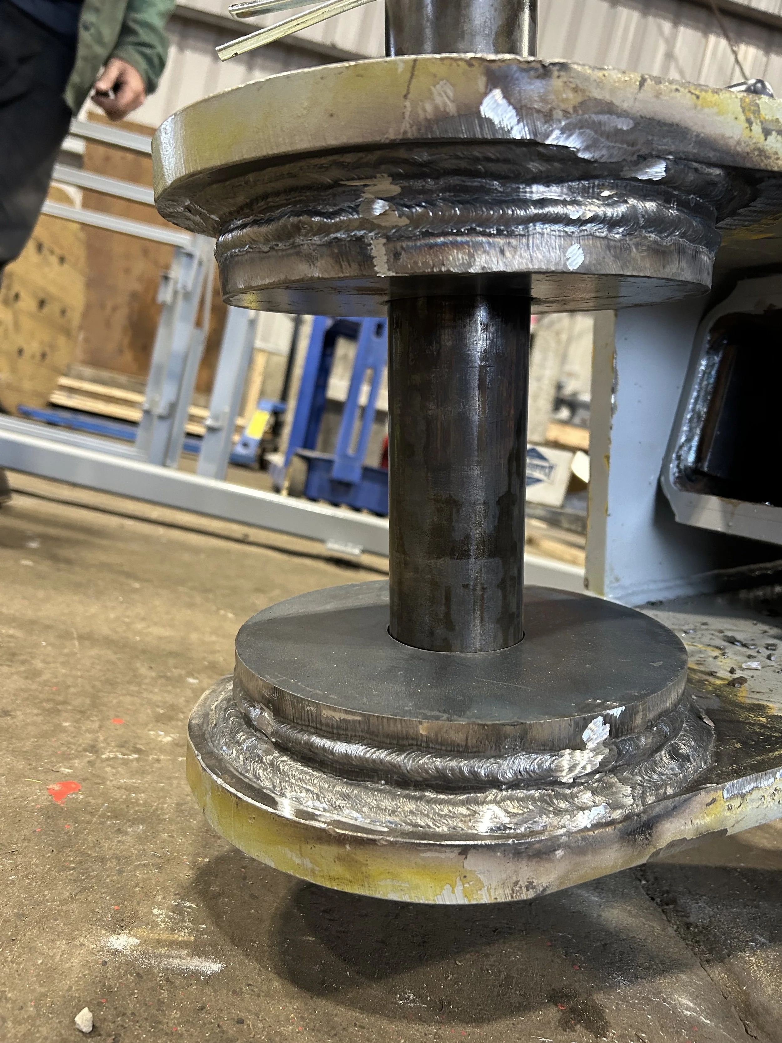 Close-up of a welded metal component with circular plates and a vertical shaft in industrial workshop setting.