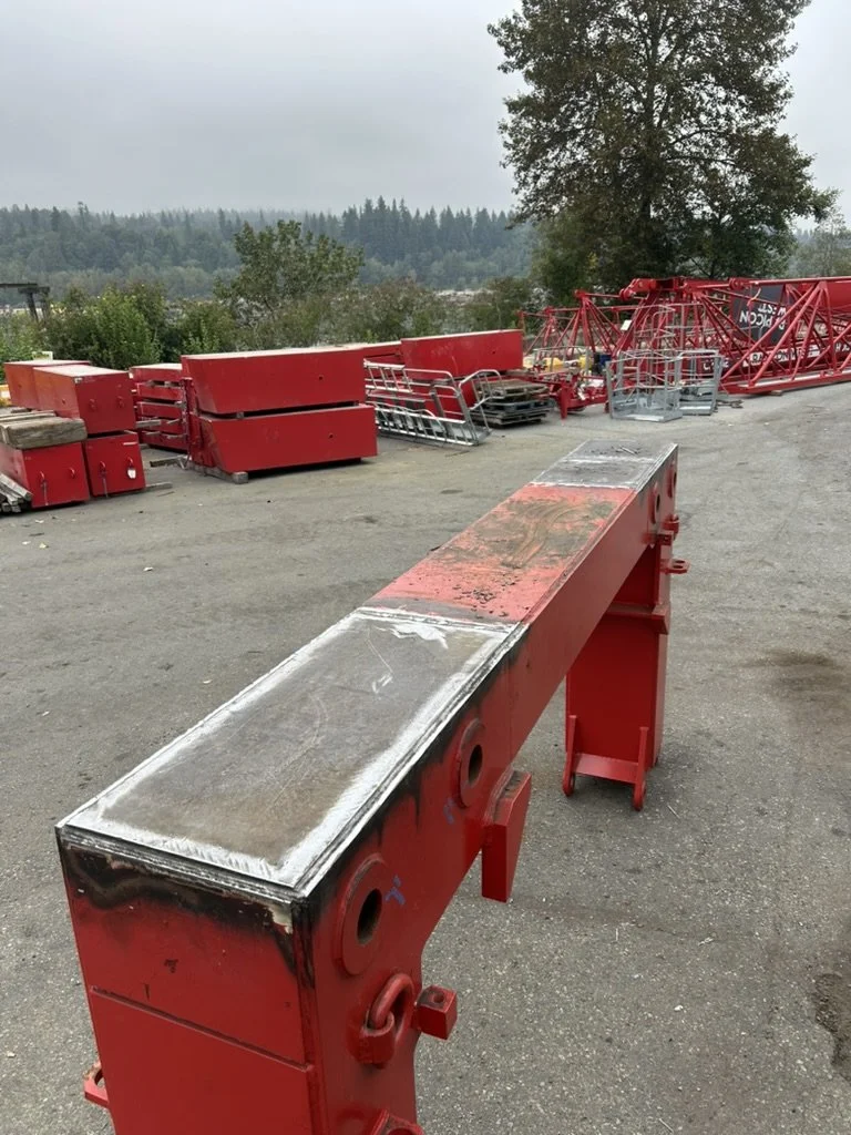 Construction site with red metal framework and equipment parts, some stacked and laid out on gray pavement, against a background of trees and overcast sky. crane component repair work
