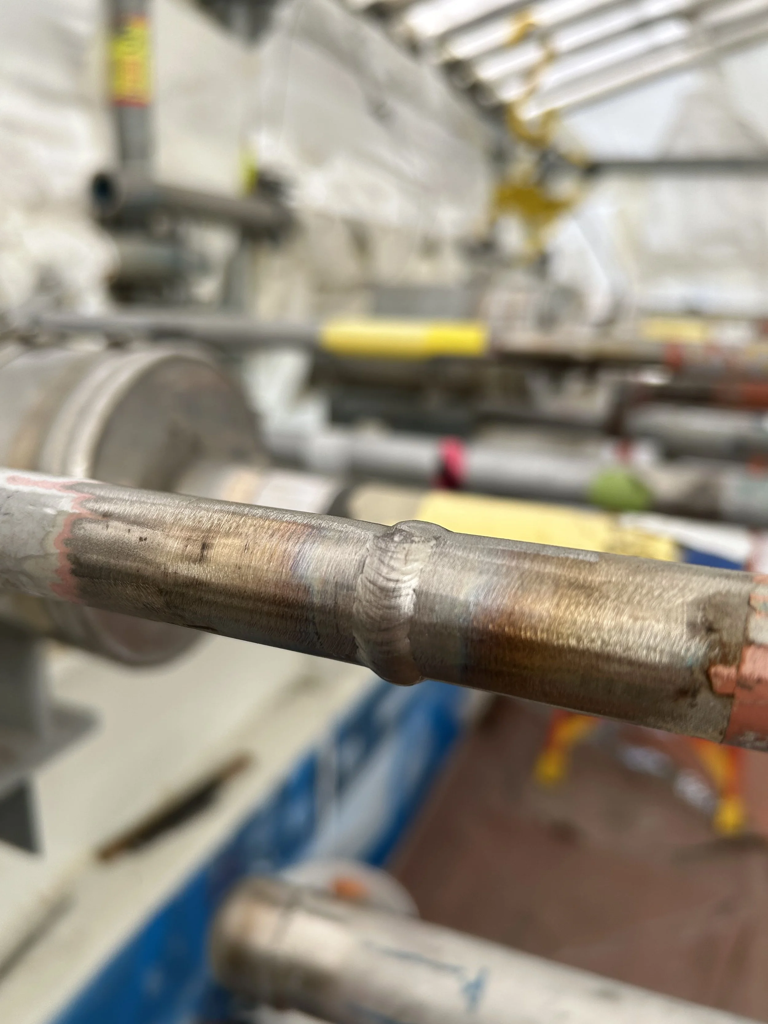 Close-up of a metal shaft with a weld bead, on an industrial machine or equipment in the background.