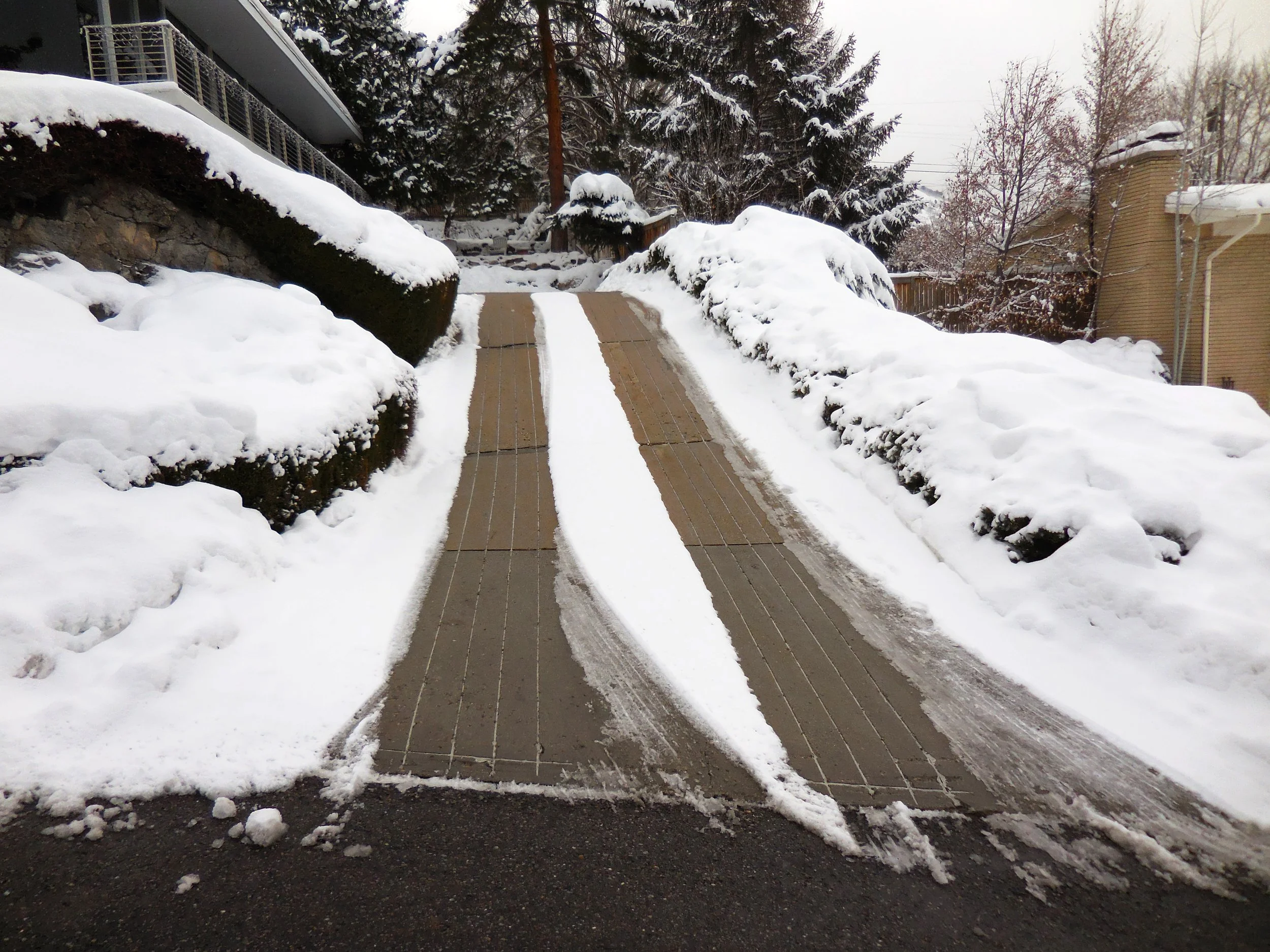 A snow-covered driveway with tire tracks, flanked by snow-covered rocks and trees, with houses and trees in the background. Driveway that does not have a carport but needs one