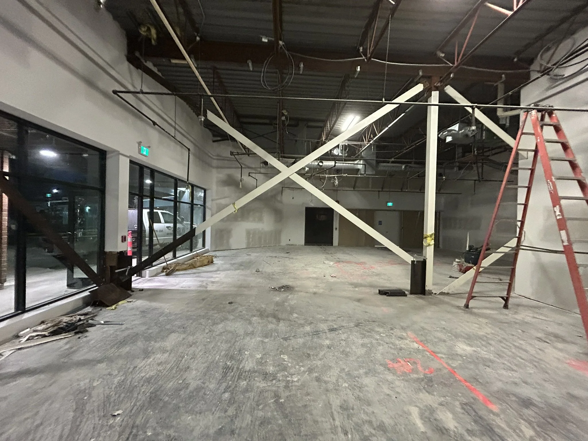 Interior construction site with metal beams and a red ladder inside a commercial building, unfinished floor, and large glass windows on the left.