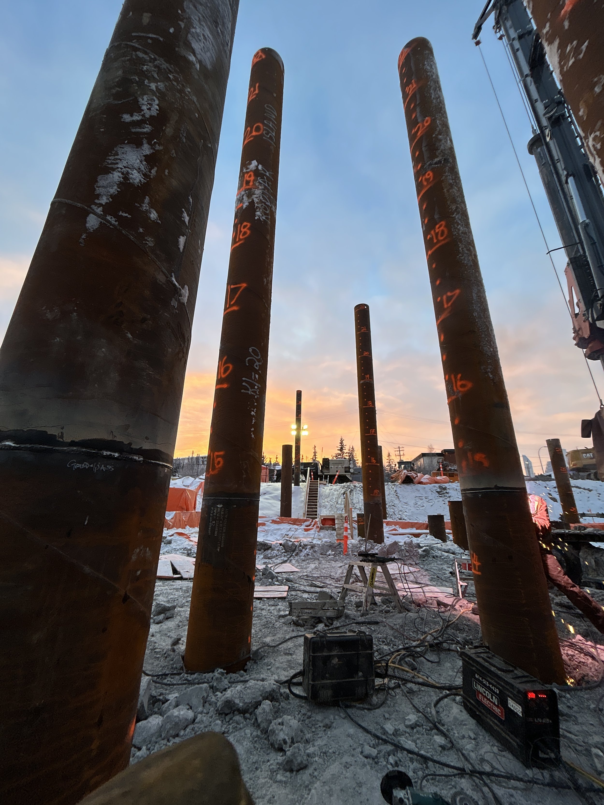 Construction site with large steel piles numbered and marked, snow on the ground, sunset in the background, construction equipment and wires visible. Pile driving construction site