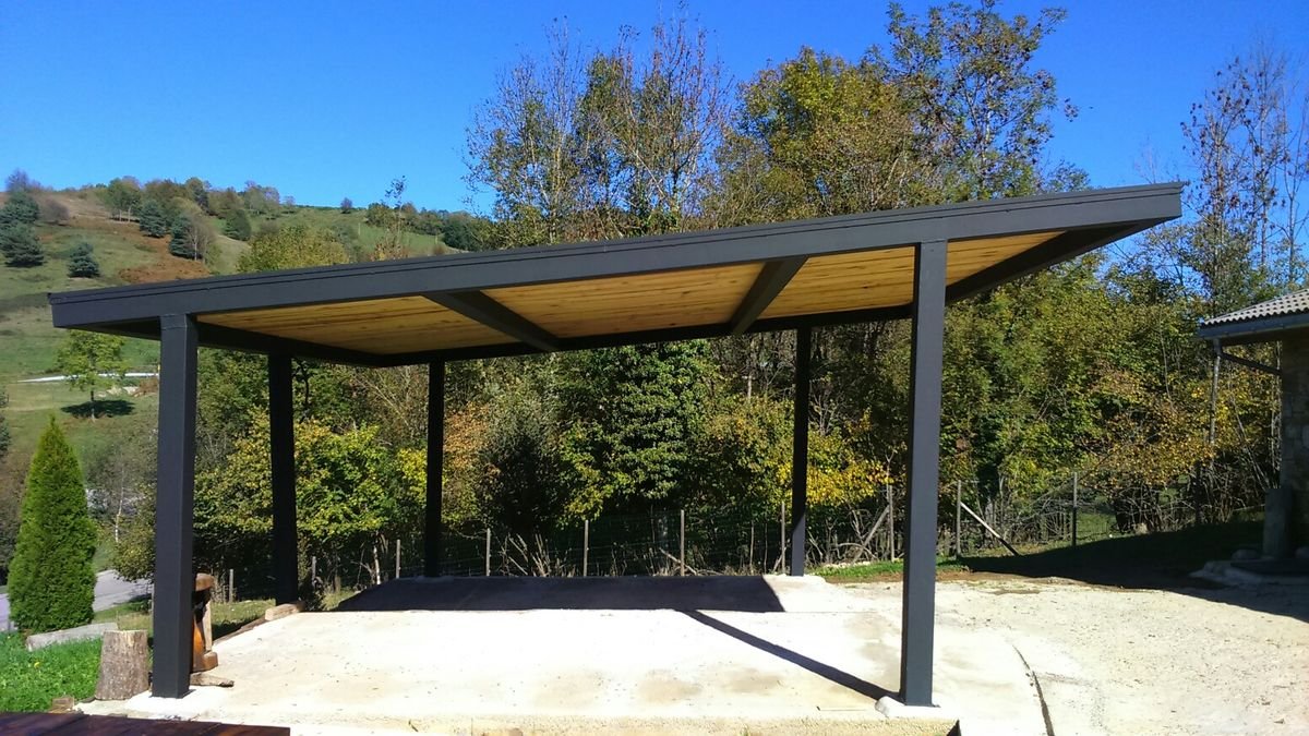 A modern carport with a black metal frame and wooden roof, situated outdoors in a rural area with trees and hills in the background.