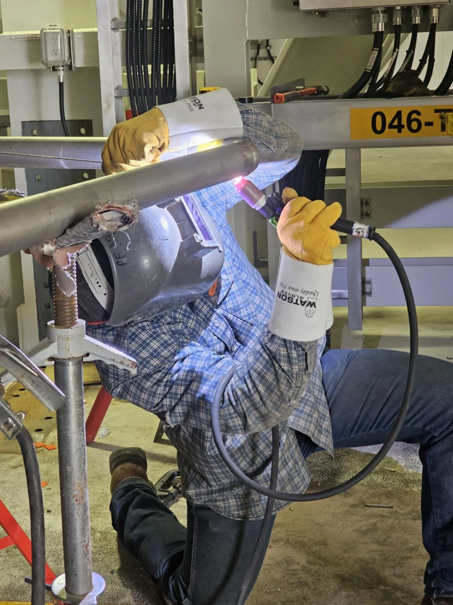 Worker welding a metal pipe while kneeling on the ground, wearing gloves and a helmet with a face shield, in an industrial setting.
