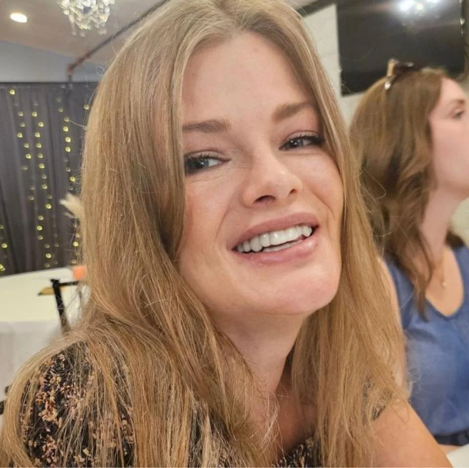 Young woman with long, wavy red hair smiling with her mouth slightly open, in an indoor setting with string lights and another woman in the background.