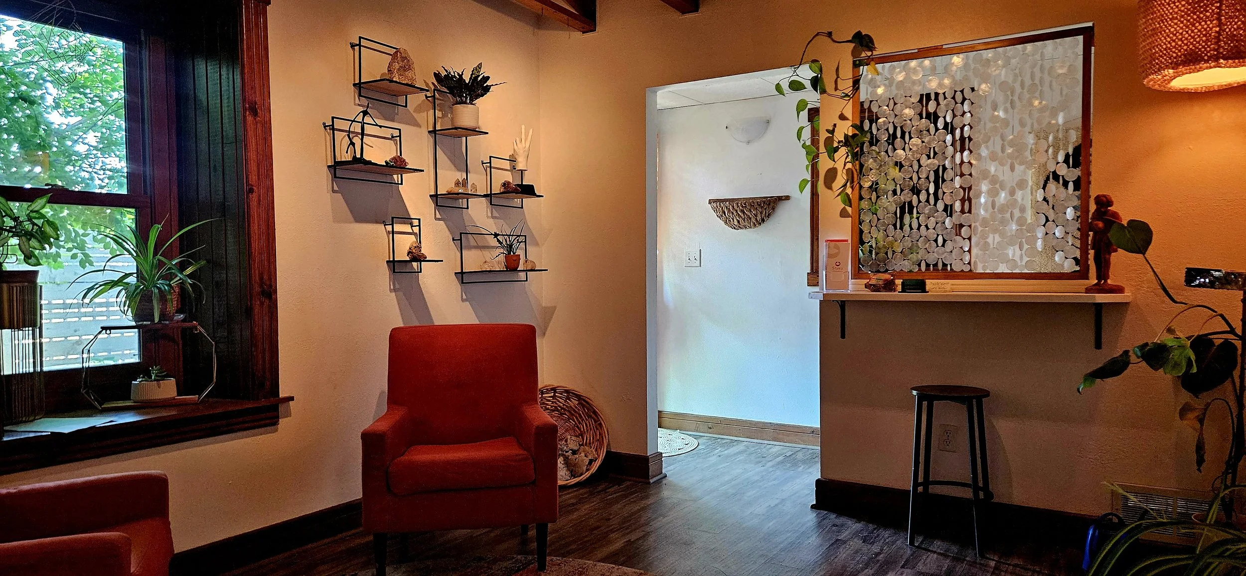 Cozy living room with orange chairs, green plants, a window with wooden frame, and decorative wall shelves with small plants and ornaments; a partition with shell decorations separates the room from an entryway.
