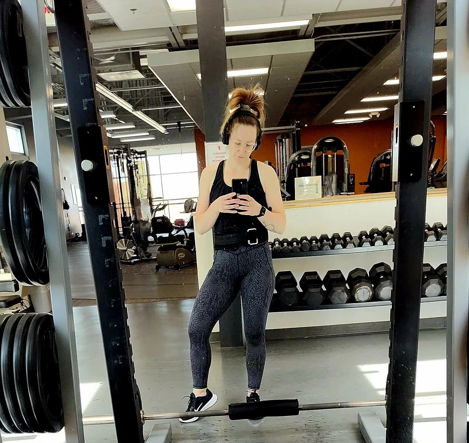 A woman taking a selfie in a gym mirror, dressed in workout clothes, standing near weight racks and exercise equipment.