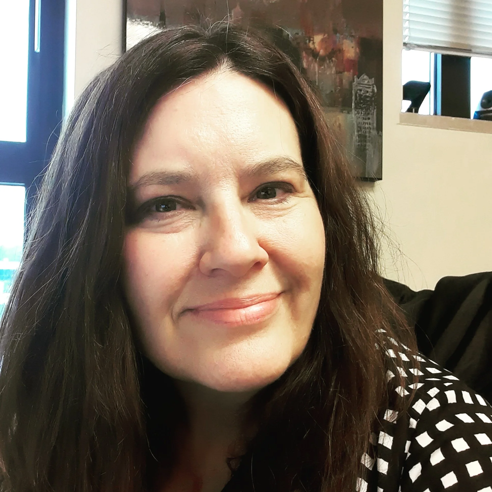 Close-up of a smiling woman with long dark hair, in an indoor setting with a painting and window blinds in the background.