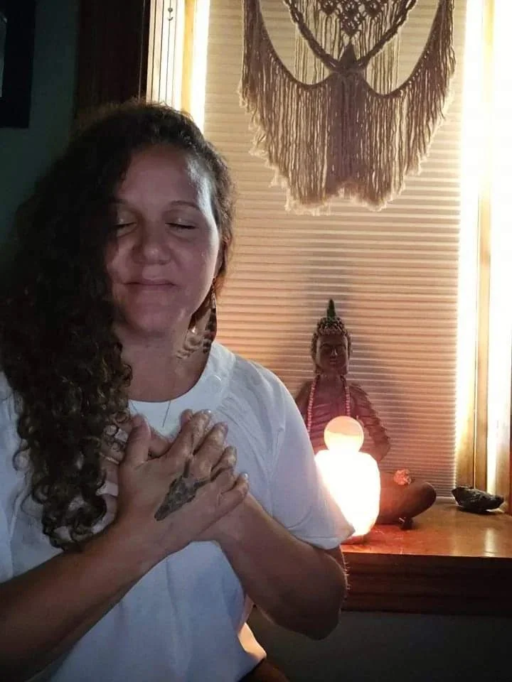 A woman with curly hair and a white shirt is smiling with her eyes closed, holding her hands on her chest, standing in front of a wooden altar with a Himalayan salt lamp and a Buddha statue, in a room with a decorative wall hanging.