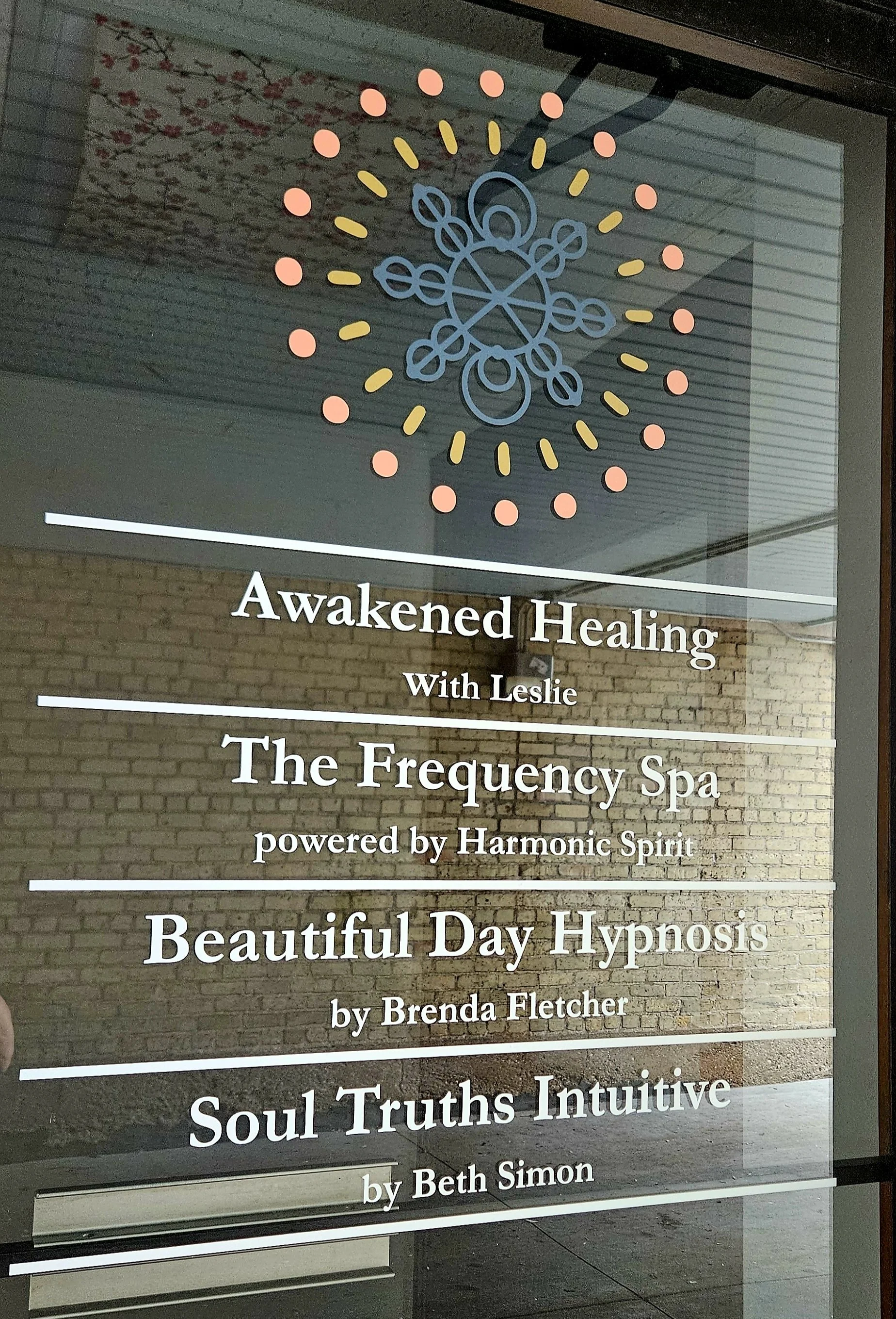 Sign for wellness and healing services featuring a geometric snowflake or mandala symbol at the top, with offerings including 'Awakened Healing with Leslie,' 'The Frequency Spa powered by Hamonic Spirit,' 'Beautiful Day Hypnosis by Brenda Fletcher,' and 'Soul Truths Intuitive by Beth Simon.' The background shows a brick wall and ceiling, with a reflection of a branch with pink blossoms on the glass.