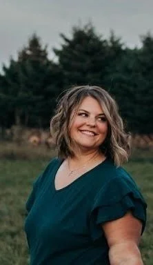 A woman with shoulder-length wavy hair smiling outdoors, wearing a dark teal top, with trees in the background.