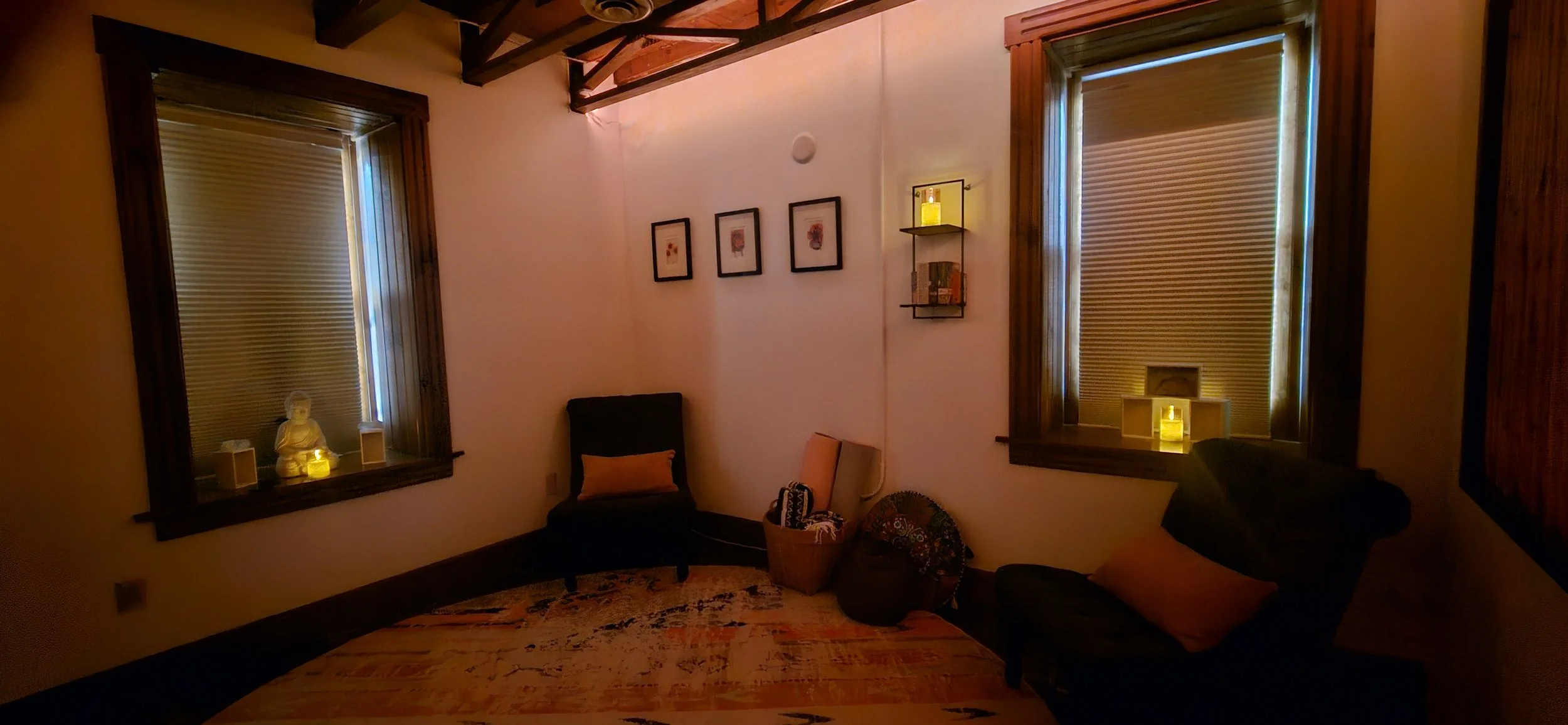 A cozy room with two windows with wooden frames and closed blinds, a small Buddha statue with candles on the windowsills, a black armchair with a cushion, a basket with items, a dark armchair with another cushion, framed pictures on the wall, and small lit candles for ambiance.