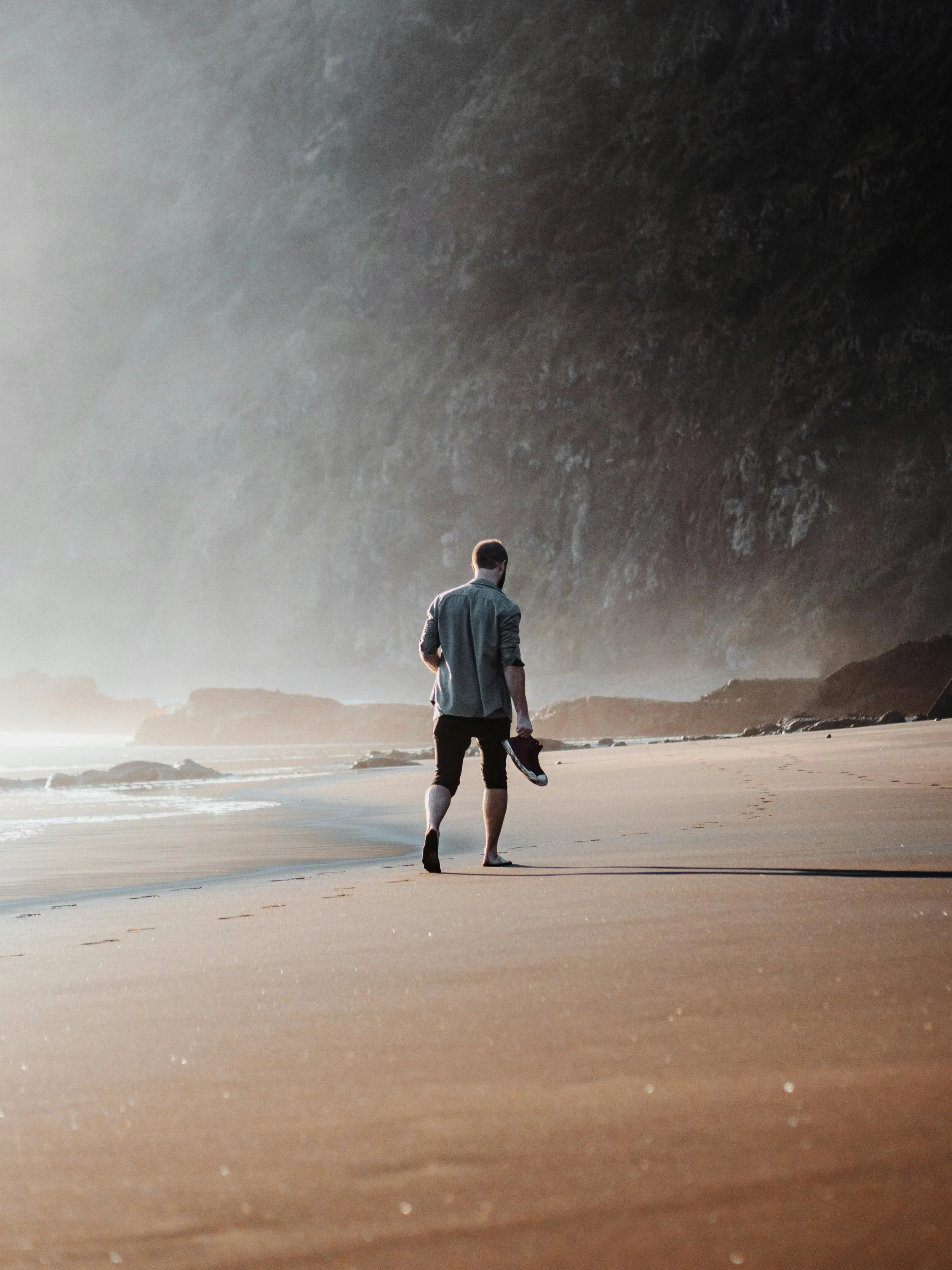 A man walking barefoot on the beach holding shoes, with cliffs and the ocean in the background.