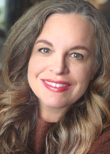 Close-up photo of a smiling woman with wavy, light brown hair and blue eyes, wearing brown clothing.