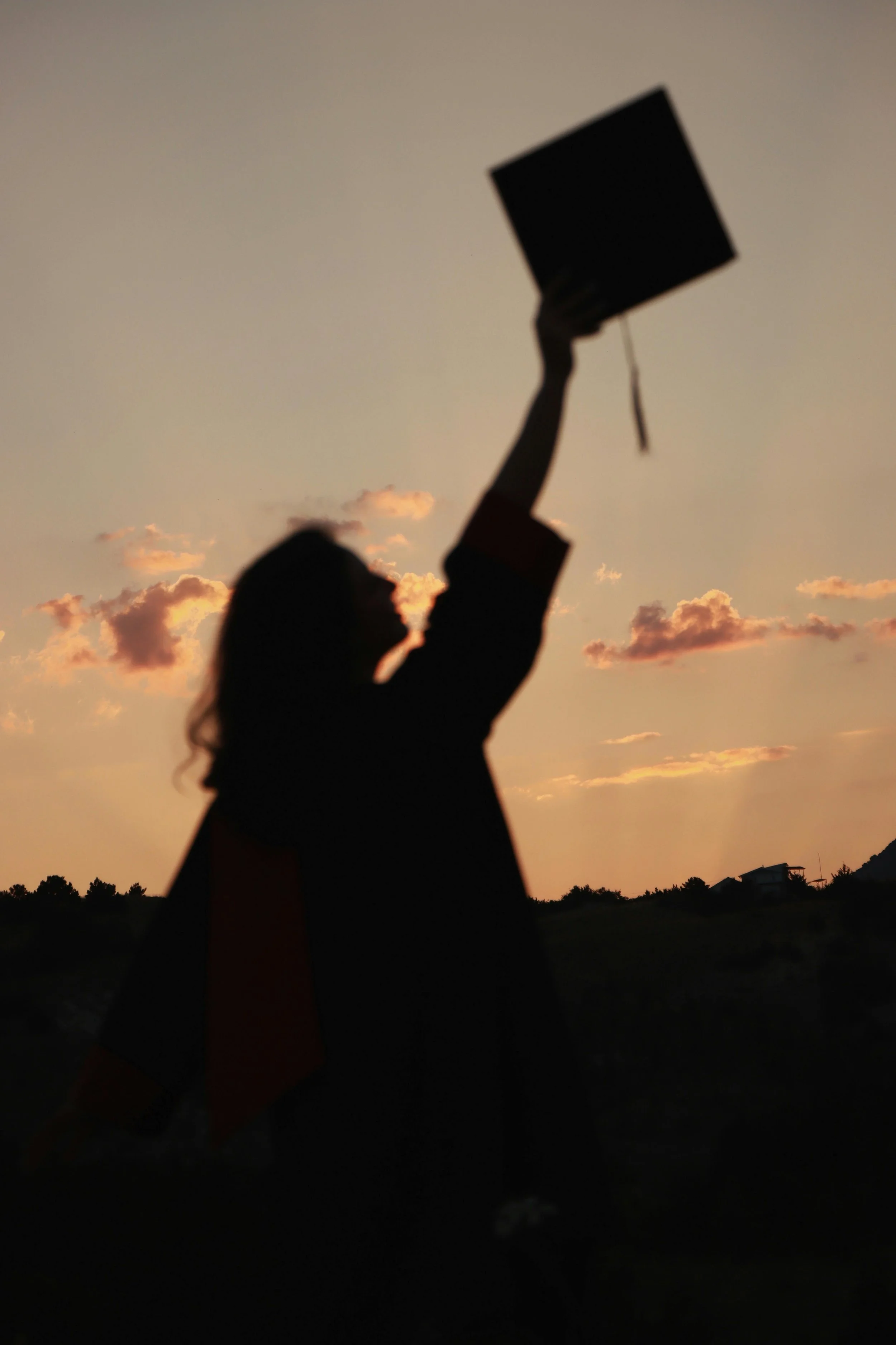 A college graduate holding her cap high at sunset.