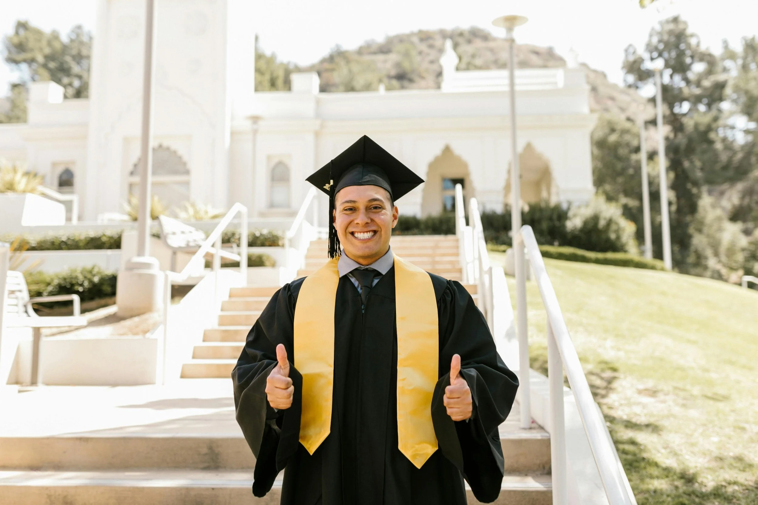 A smiling young man in a graduation cap and gown standing outdoors in front of a white building, giving thumbs up.