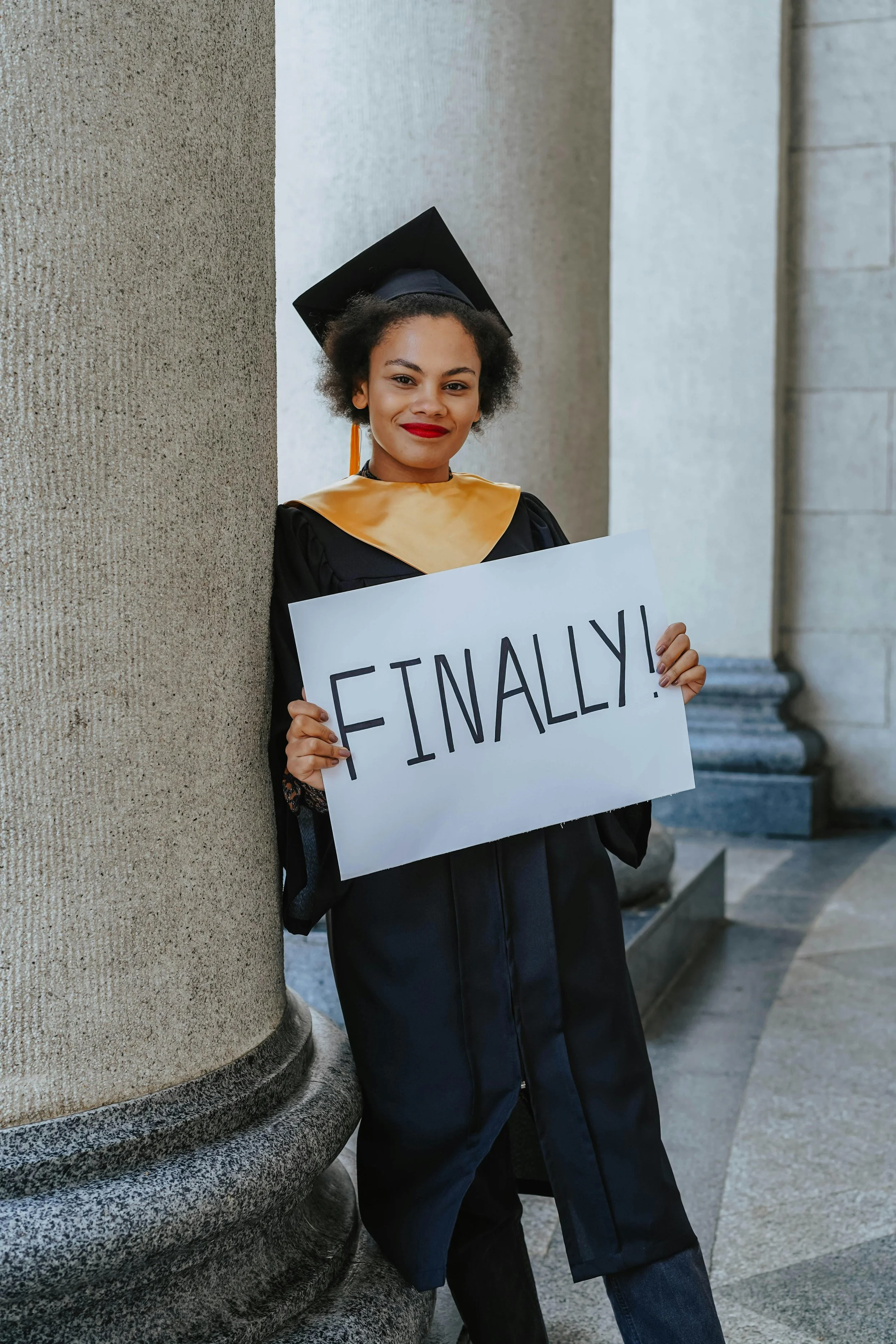 Young woman in graduation gown and cap holding a sign that says 'Finally!' with a smile, standing against a pillar in an indoor setting.
