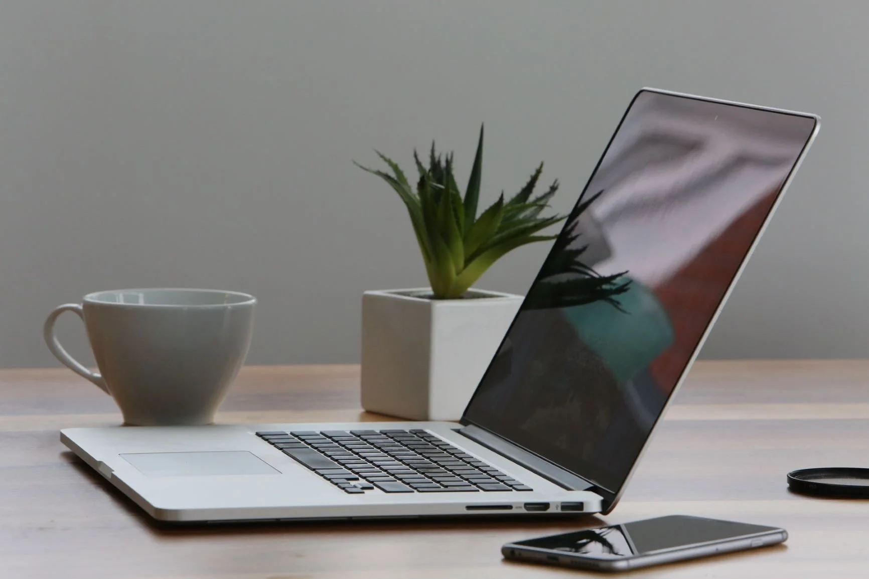 Open silver laptop on a wooden desk, with a white coffee mug, a potted green plant, and a smartphone nearby.