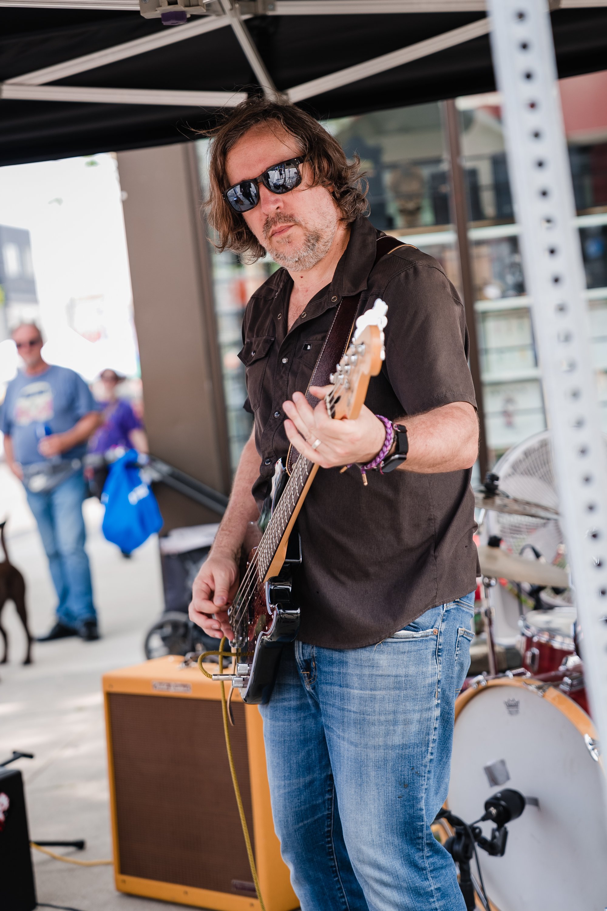 A man with shoulder-length brown hair and sunglasses playing an electric guitar at an outdoor event, with onlookers and a drum set in the background.