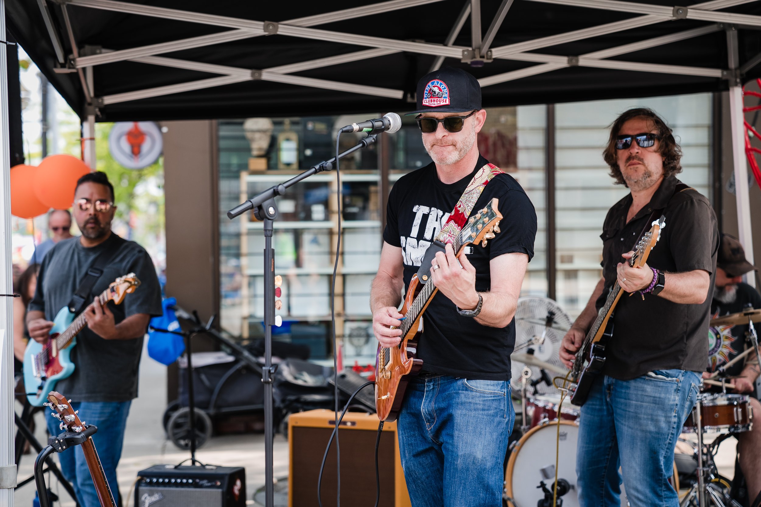 A band performs on an outdoor stage under a black canopy, including a man in a black t-shirt, sunglasses, and a black cap with a red and blue patch, playing an electric guitar. Other band members play guitars and drums in the background.