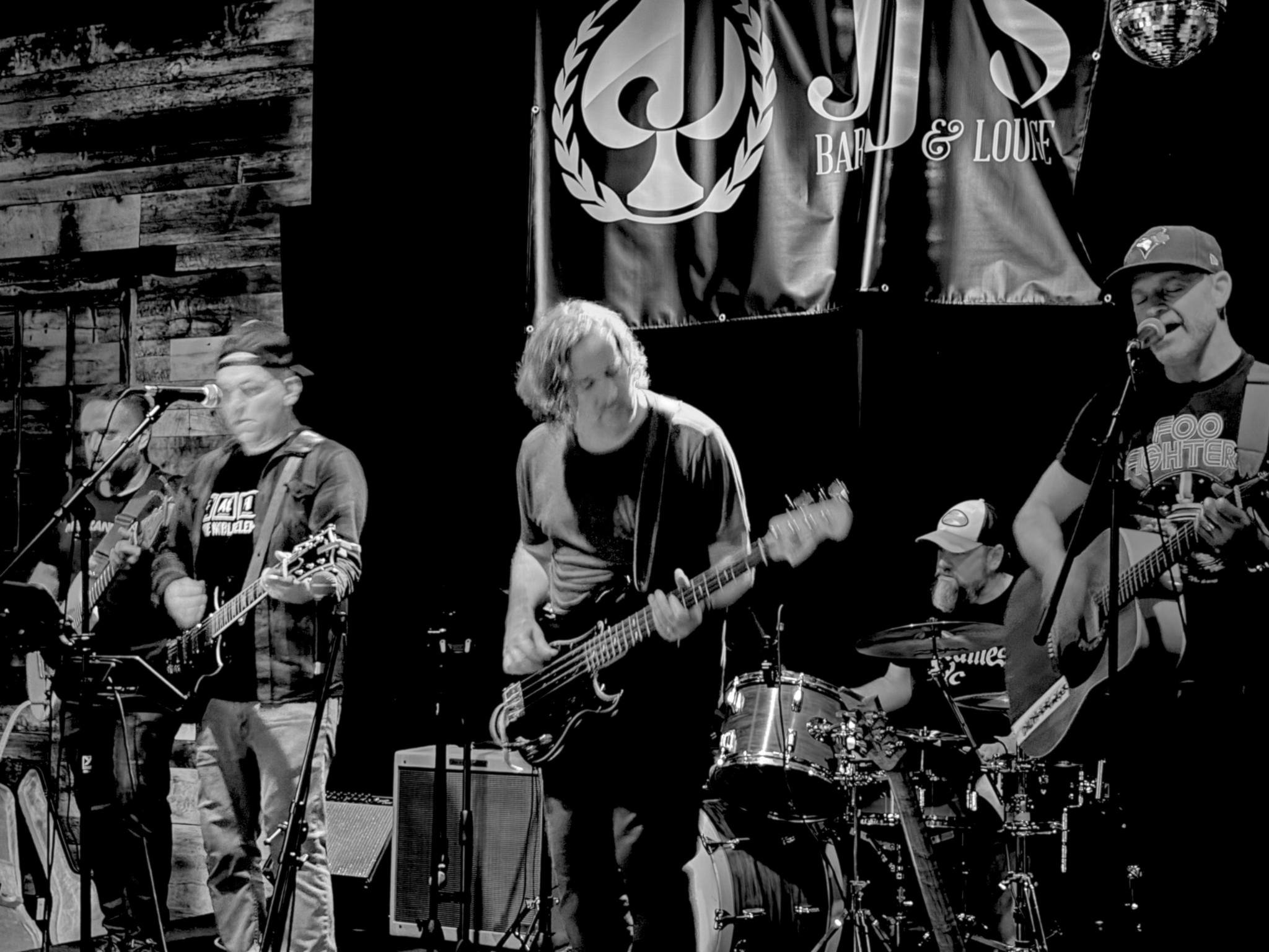 Black and white photo of a band performing on stage with four guitarists, a drummer, and a vocalist, in a bar and lounge environment with wooden walls and a large banner.