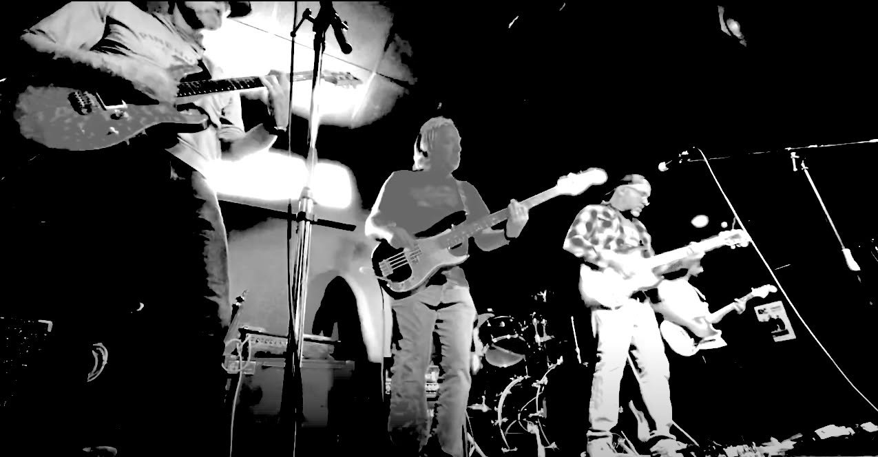 Black and white photo of a four-member band performing on stage with guitars and drums, stage lighting illuminating the musicians.