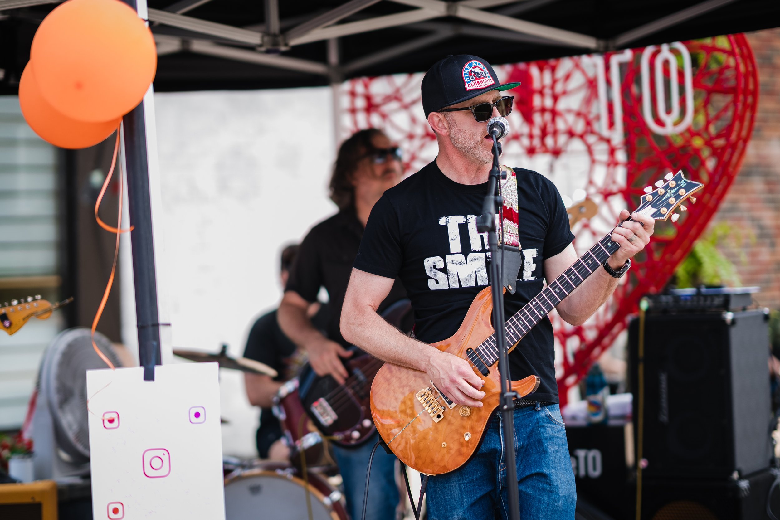 Man playing electric guitar and singing into a microphone at an outdoor event, wearing sunglasses and a black t-shirt. Two musicians are visible in the background, one playing bass guitar. There are orange balloons and social media icons on a sign in