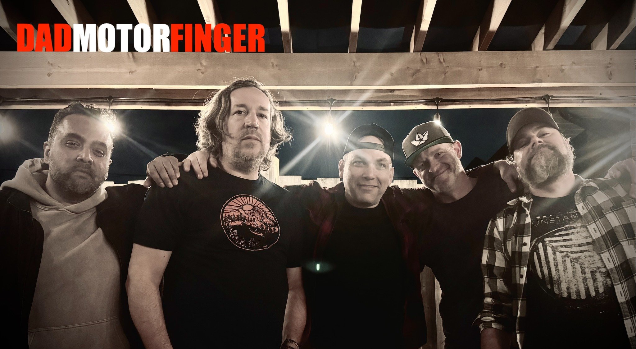 A group of five men standing close together outdoors at night, with a wooden roof overhead and bright lights behind them. The text "DAD MOTORFINGER" is displayed in the top left corner.