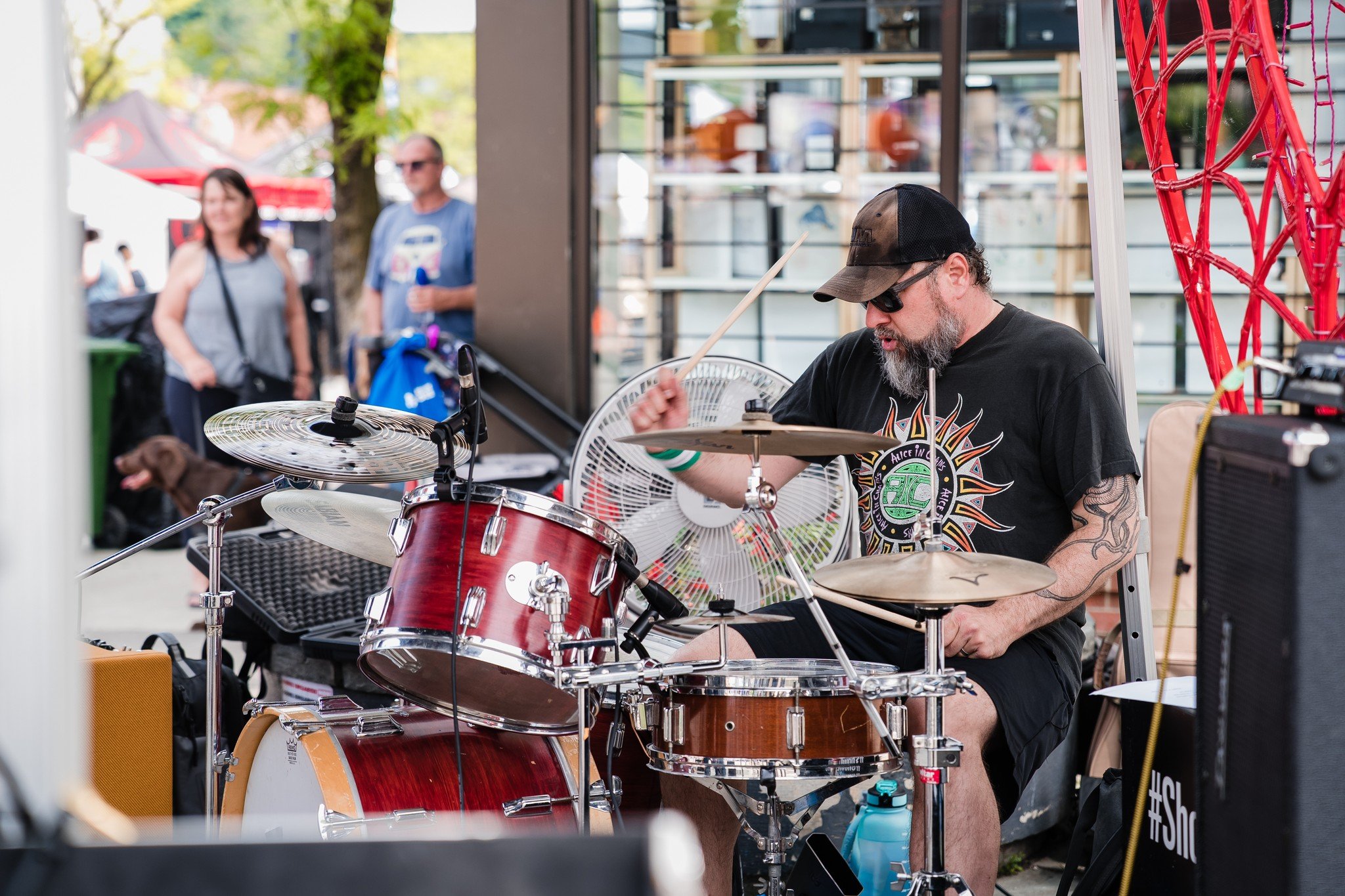 A man wearing a black t-shirt, sunglasses, and a baseball cap playing a red drum set outdoors, with people walking and standing in the background.