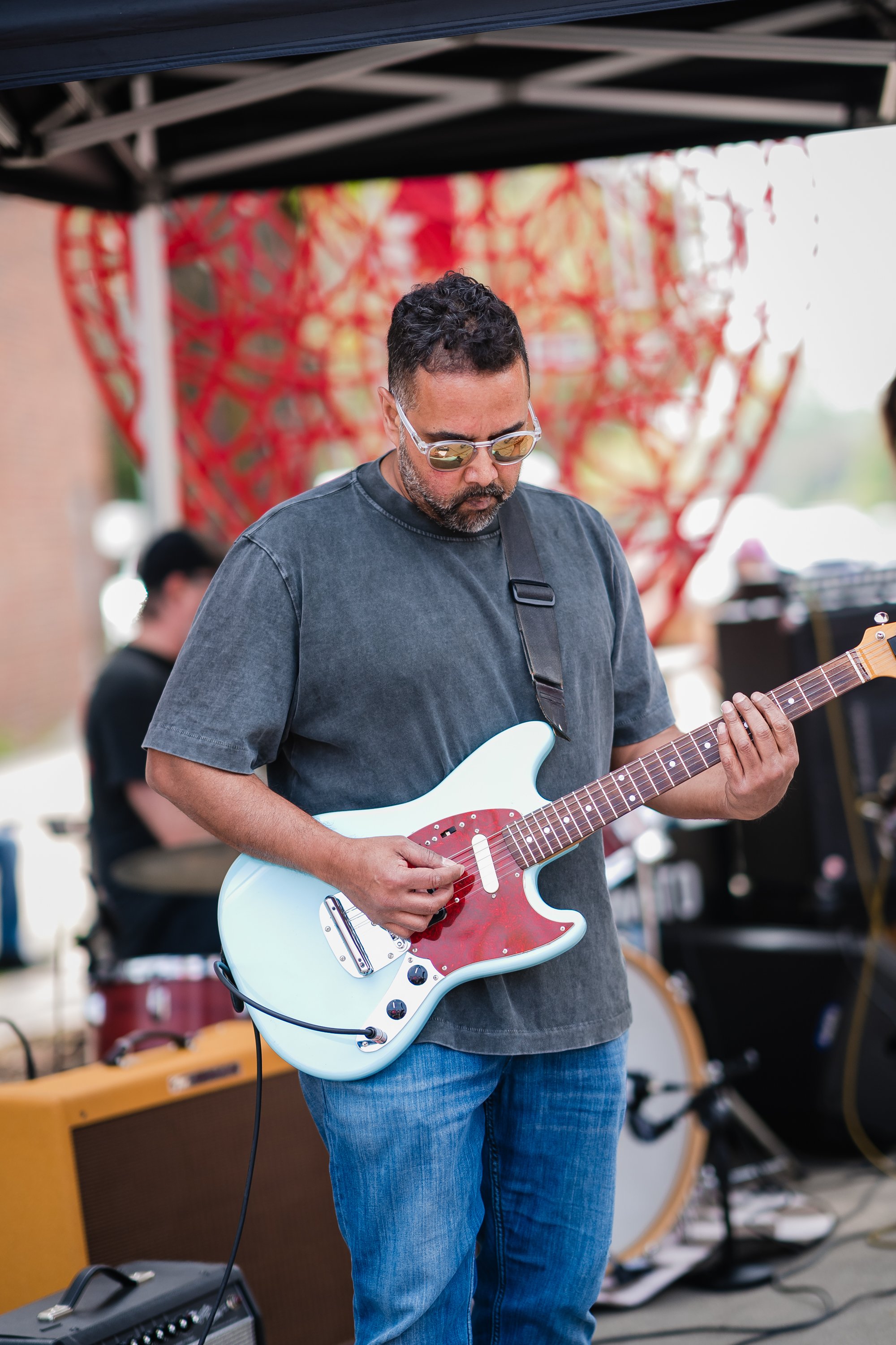 A man with curly dark hair, wearing sunglasses, a dark T-shirt, and blue jeans, playing a white electric guitar outdoors during a live music performance. There is a colorful fabric backdrop and other band members in the background.