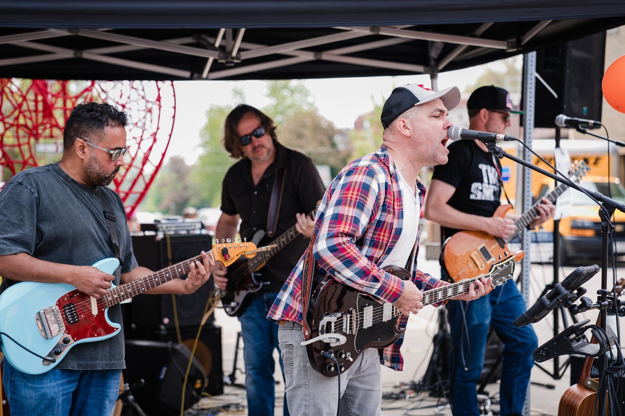 A band performing outdoors under a black tent, with four members playing guitars and singing into microphones.