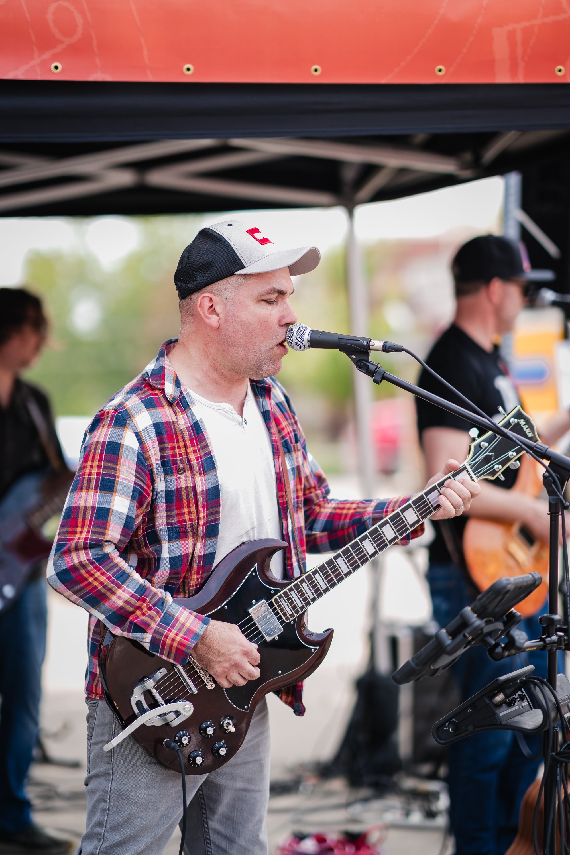 A man wearing a baseball cap and plaid shirt plays electric guitar and sings into a microphone at an outdoor music event, with other musicians in the background under a canopy.