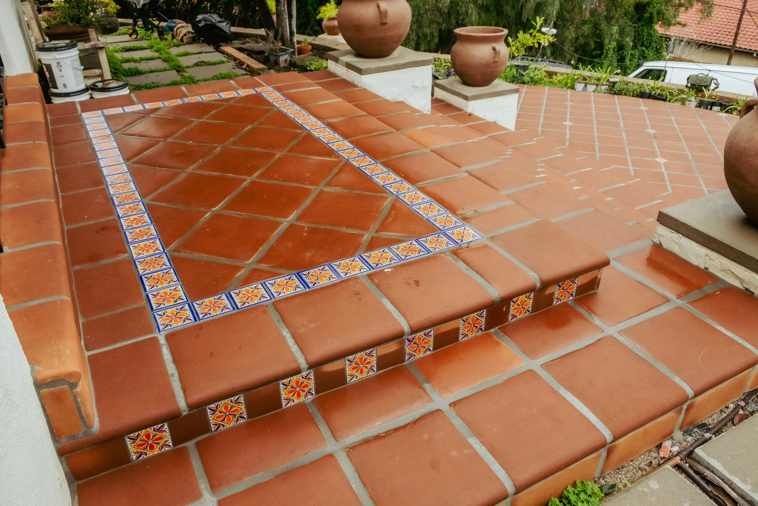 Patio with terracotta tiles and decorative tile border, featuring potted plants and a garden in the background.