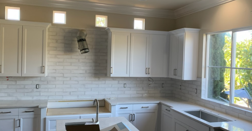 Kitchen with white cabinets, a gray brick backsplash, and a large window overlooking greenery, with some ceiling and wall fixtures and outlets visible.