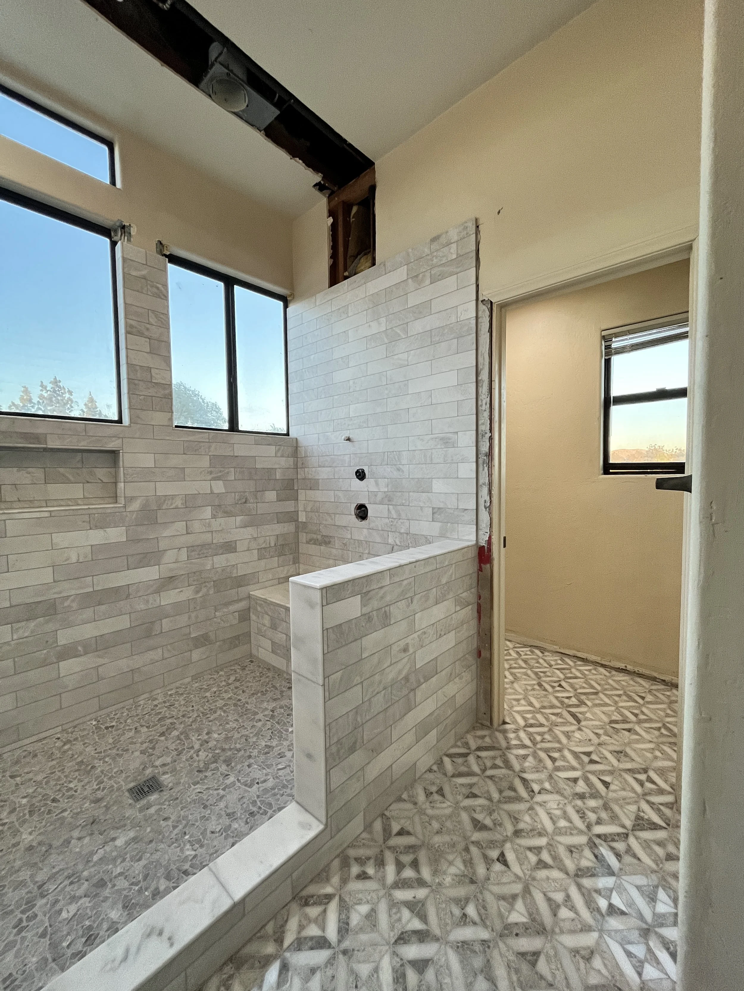 Bathroom under renovation with a partial shower area featuring beige tile walls, a pebble floor, and a partial view of a toilet in the adjacent room, with stone geometric patterned flooring and windows providing natural light.