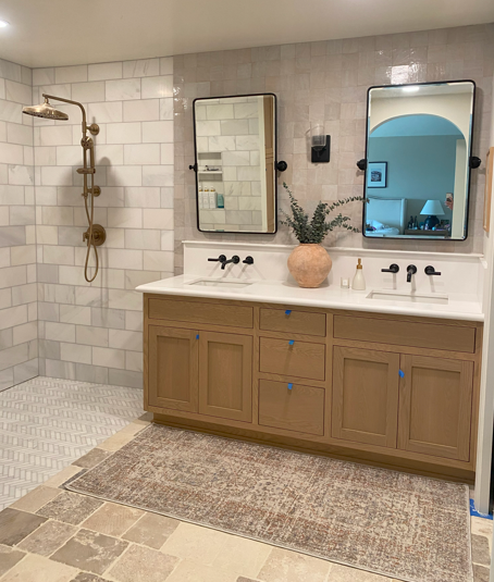 Bathroom with a double vanity, large mirrors, a potted plant, and a walk-in shower with a gold shower system.
