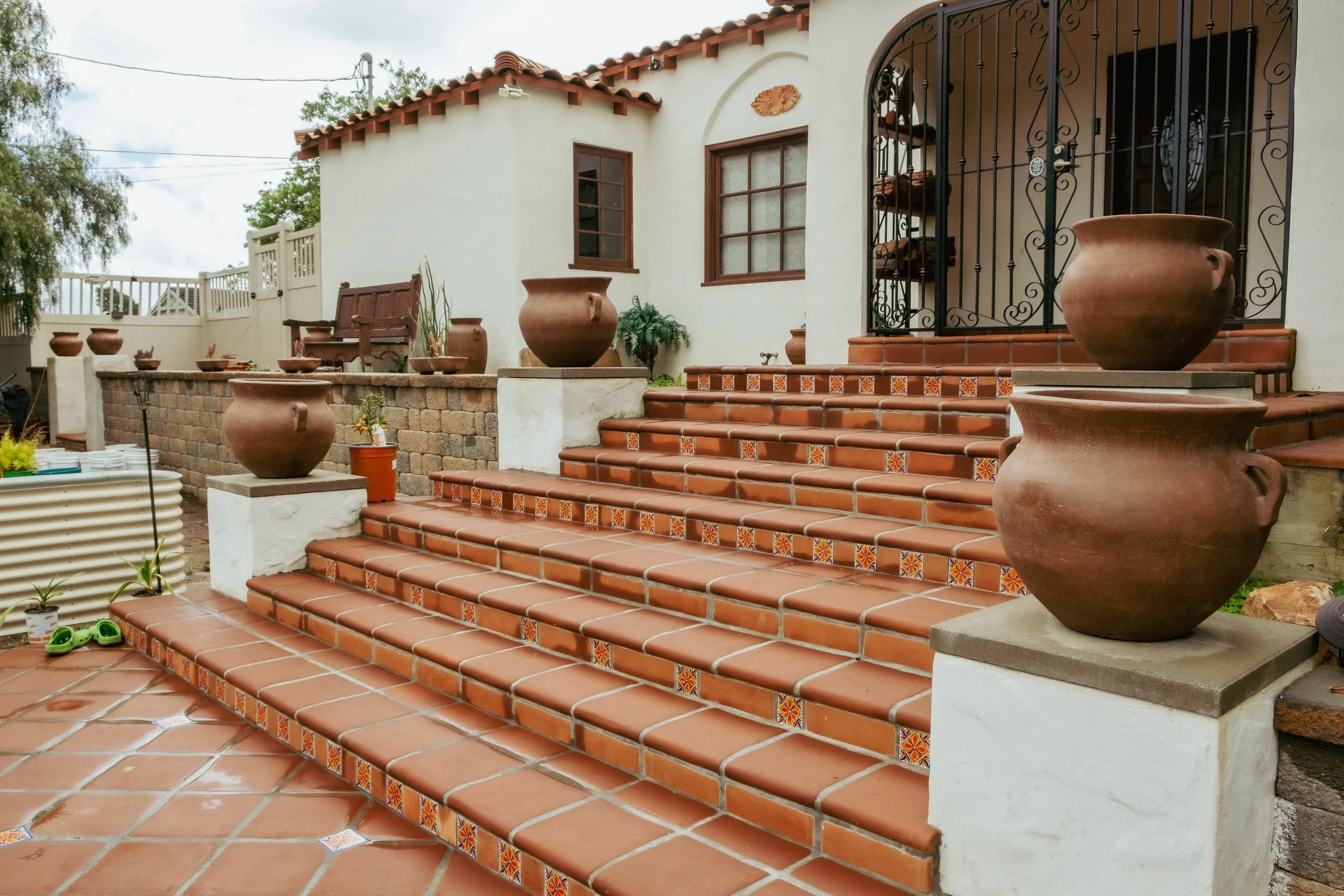 Terrace with red tile stairs, large clay pots on pedestals, decorative tiles on stairs, white stucco house with windows and iron gate, potted plants, and wooden bench.