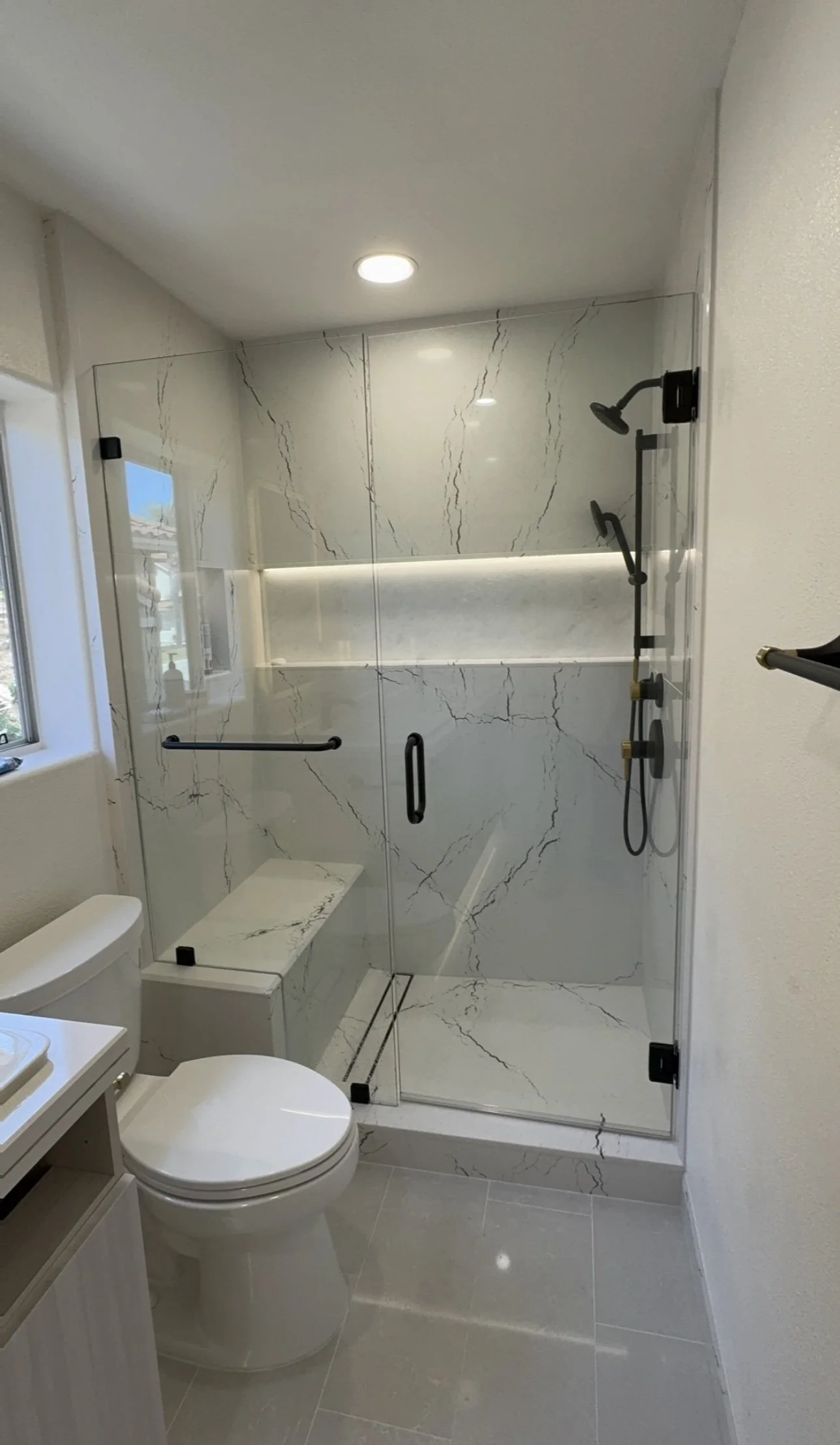 Bathroom with a glass-enclosed shower featuring white and gray marble walls, a built-in marble bench, and black shower fixtures. There is a frosted window and beige tile flooring.