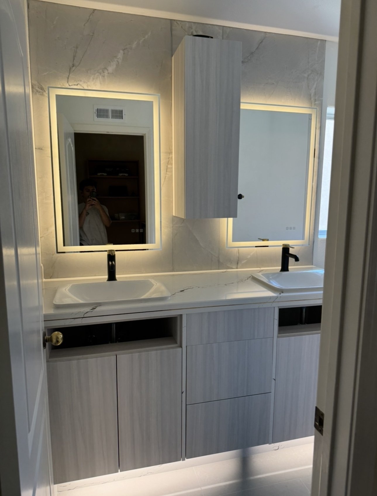 Modern bathroom with two illuminated mirrors, dual sinks, and light wood cabinetry.