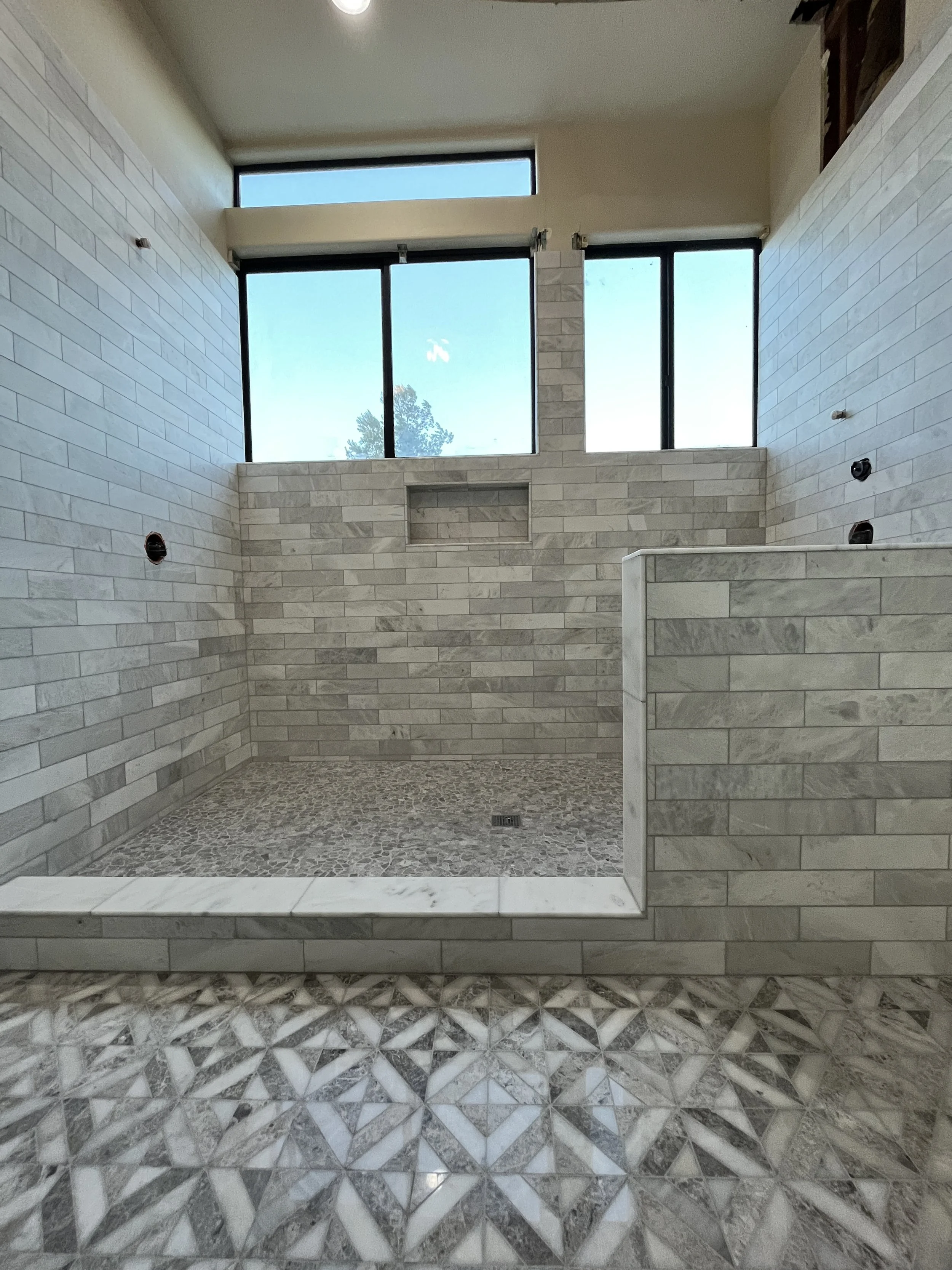 Empty walk-in shower with gray subway tile walls, pebble tile floor, and window with natural light.