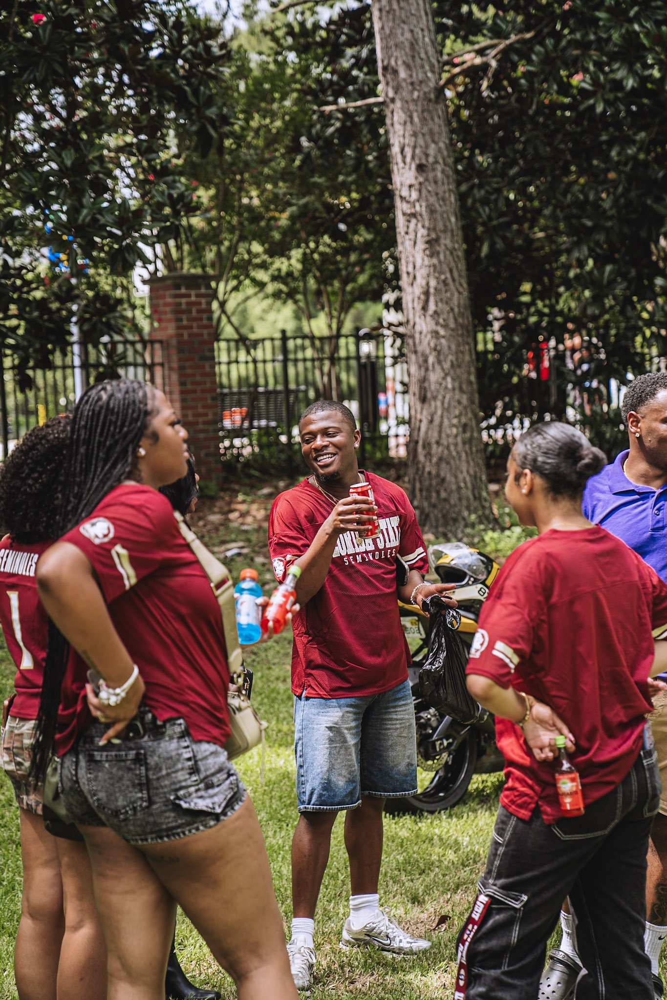 A group of people gathered outdoors, some wearing FSU football jerseys, talking and smiling. One man in the center is holding a drink and smiling.