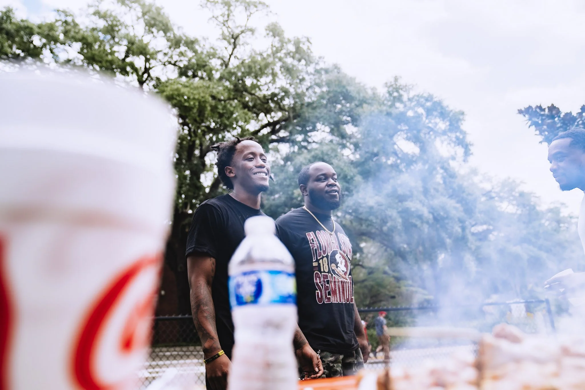 Two men smiling at a barbecue grill outdoors, with smoke and trees in the background, and bottles and food in the foreground.