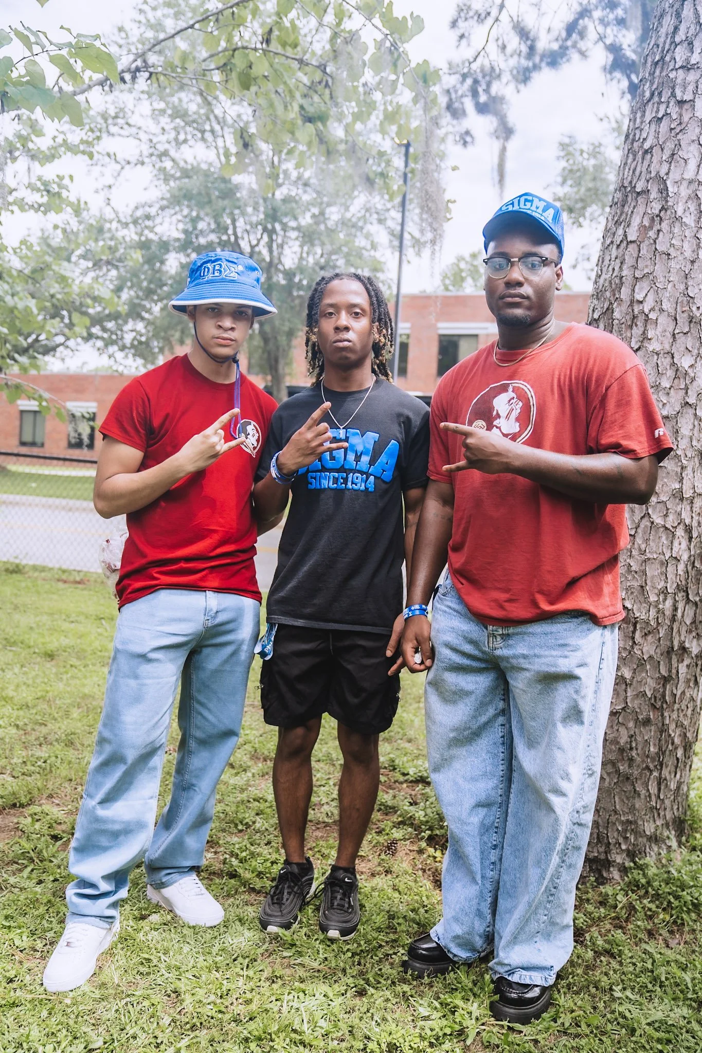 Three young men standing outdoors near a tree, making hand signs and wearing casual clothing with fraternity symbols.