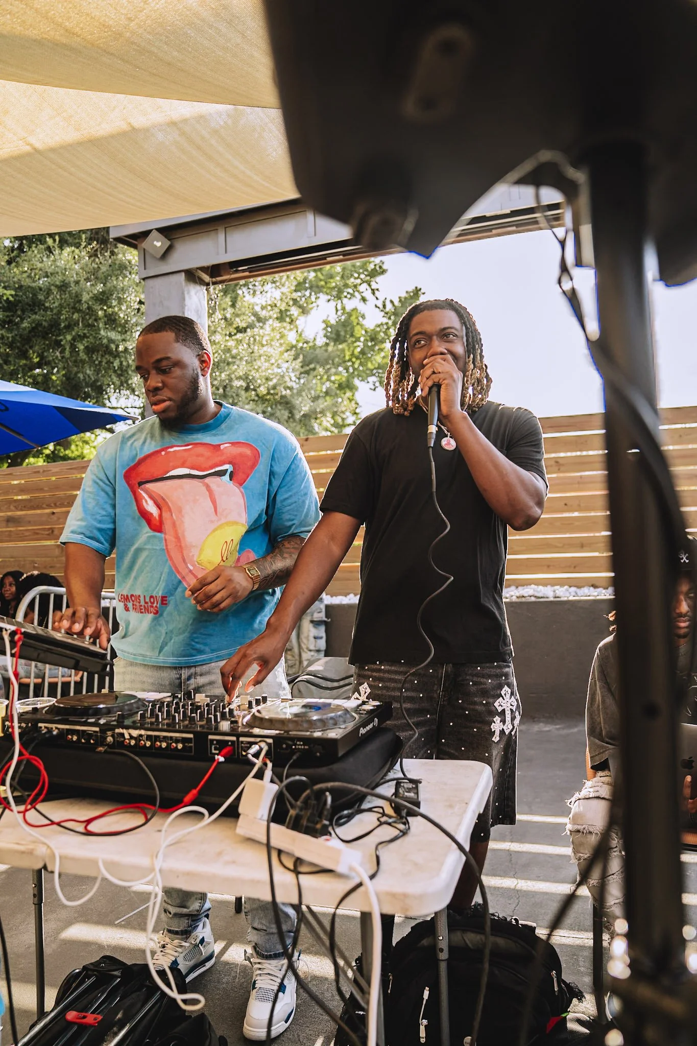 Two men performing at an outdoor event with DJ equipment, one singing into a microphone and the other adjusting the DJ controller, with a wooden fence and green trees in the background.